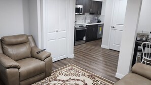 A modern living room with a leather armchair, a patterned rug, and a kitchen area with dark cabinets and a microwave. - CNEORM HAVEN (Edmonton)
