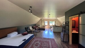 A spacious loft bedroom with a large bed, a sitting area with chairs, a kitchen area, and a view of the outdoors.