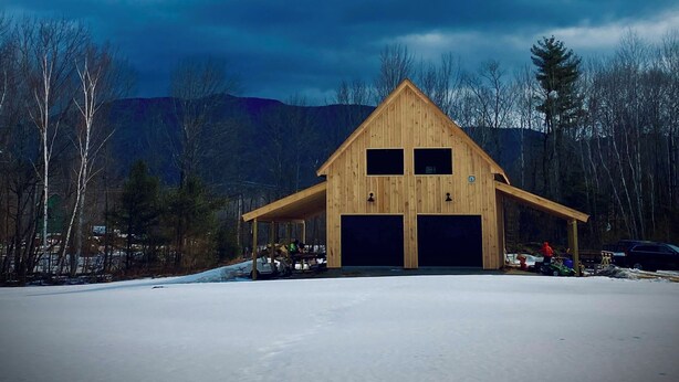 A wooden barn with two garage doors, surrounded by a snowy landscape and trees.