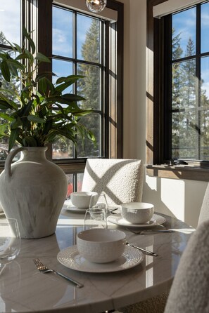 Uma área de refeições com uma mesa posta para dois, um vaso grande de plantas verdes e uma janela com vista para árvores.
