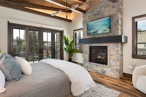 A bedroom with a stone fireplace, a large bed, and a view through a sliding glass door. - Riverwalk Luxury Home | Hot Tub + Fireplace | Walk to Trails (Winter Park)