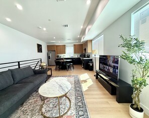 Una sala moderna con un sofá, una mesa de centro redonda y un televisor. Se observa una cocina al fondo con armarios de madera y un refrigerador.