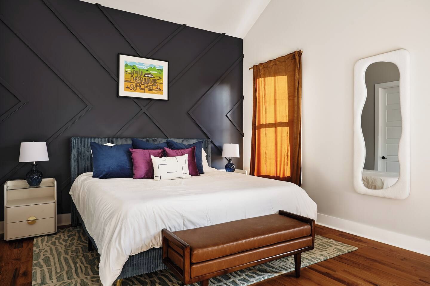 Un dormitorio con cama, mesitas de noche, un banco, un espejo en la pared y una ventana con cortinas.