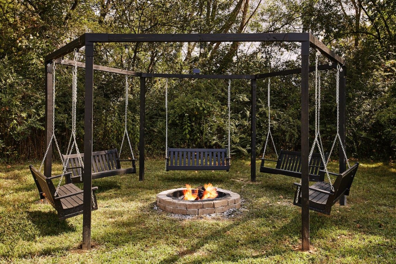 A wooden swing set with benches, a central fire pit, and a grassy area.