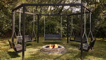 A wooden swing set with benches, a central fire pit, and a grassy area.