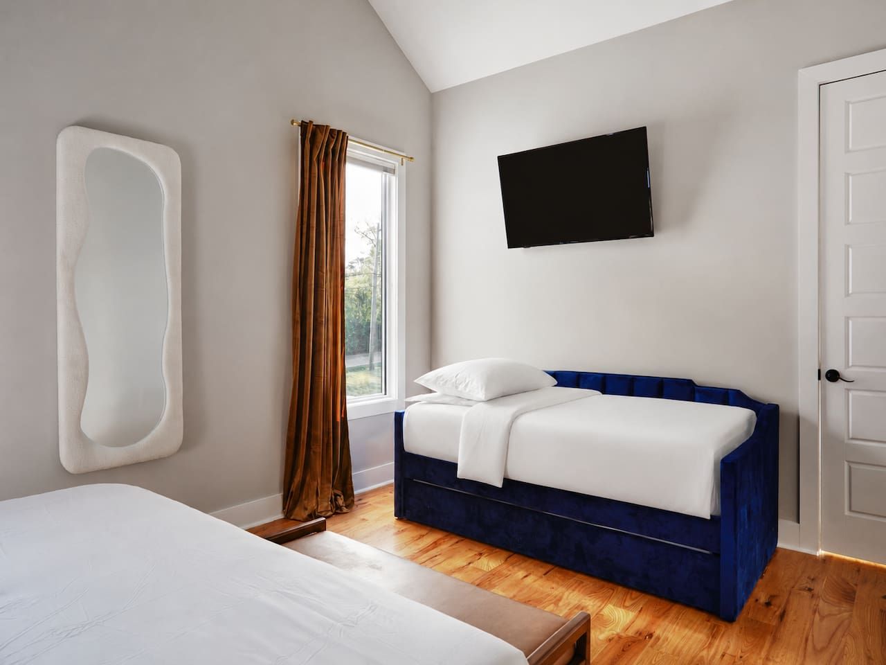 Ein Schlafzimmer mit einem Bett, einem blauen, gepolsterten Kopfteil, einem Wandfernseher und einem Fenster mit Vorhängen.