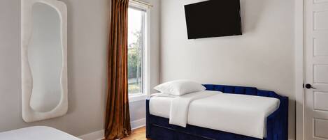 Ein Schlafzimmer mit einem Bett, einem blauen, gepolsterten Kopfteil, einem Wandfernseher und einem Fenster mit Vorhängen.