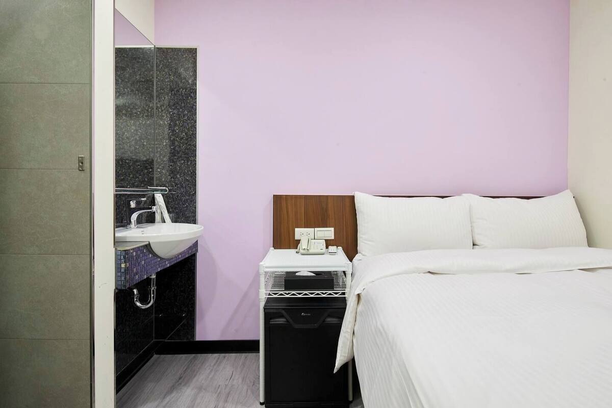 A hotel room with a bed, a bedside table with a phone and a lamp, a bathroom with a sink and mirror, and a wooden headboard.