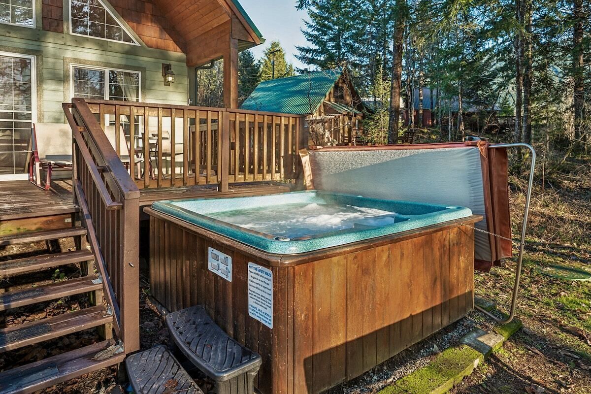 Un bain à remous en bois, agrémenté d’un panneau de sécurité, une terrasse en bois et un escalier menant à une maison entourée d’arbres.