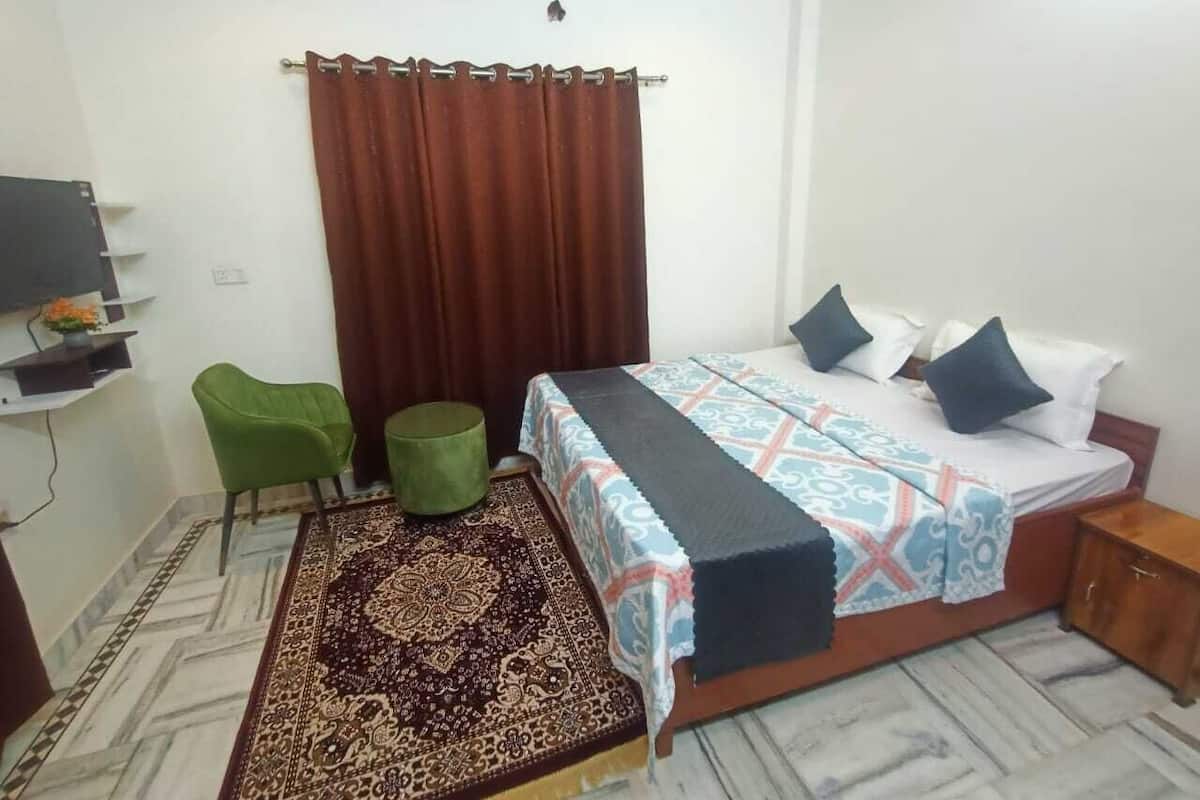 Un dormitorio con cama, una silla verde, un taburete pequeño verde y una alfombra estampada.