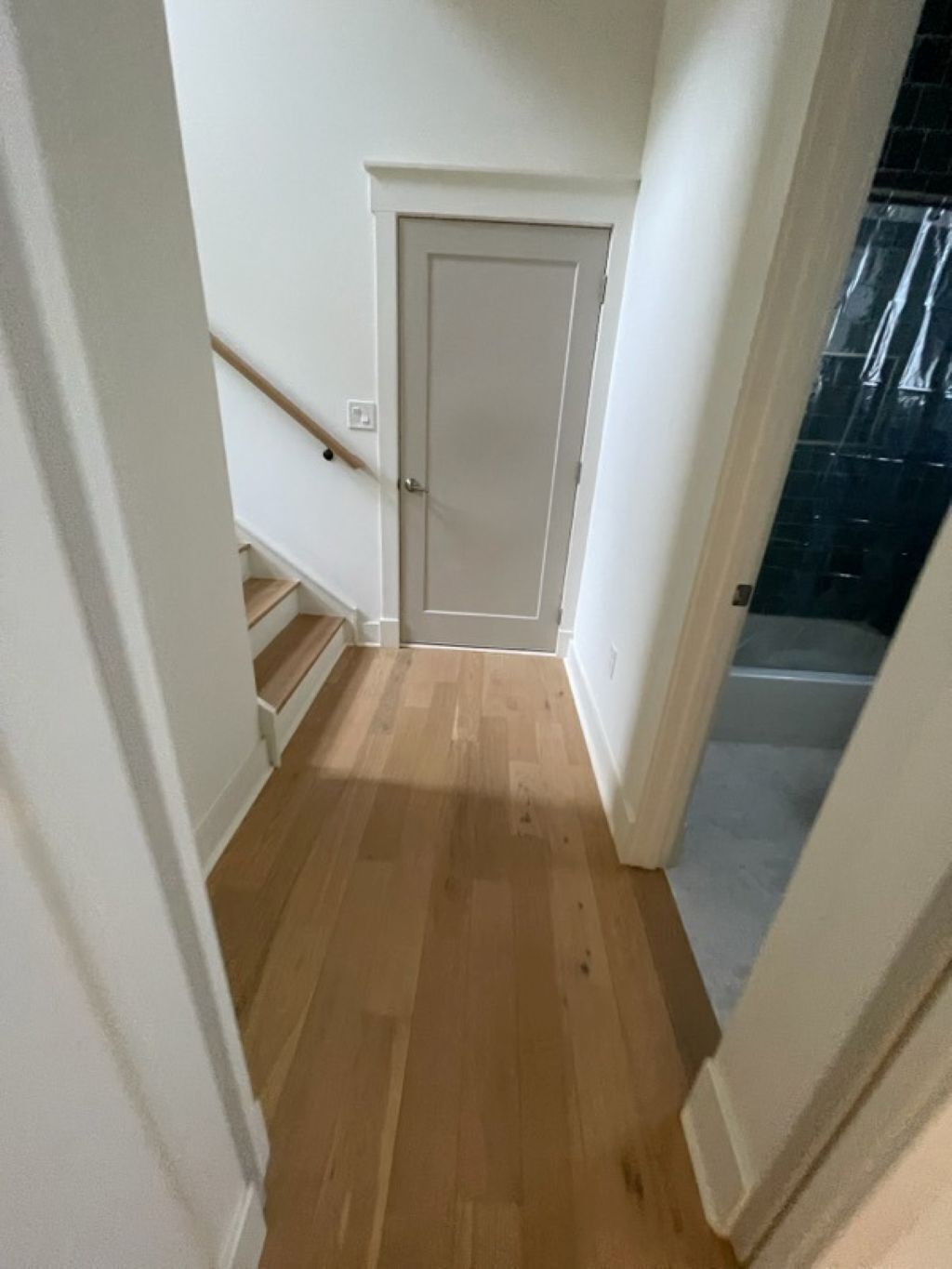 A narrow hallway with wooden flooring, a white door, and a staircase leading to a black tiled area.