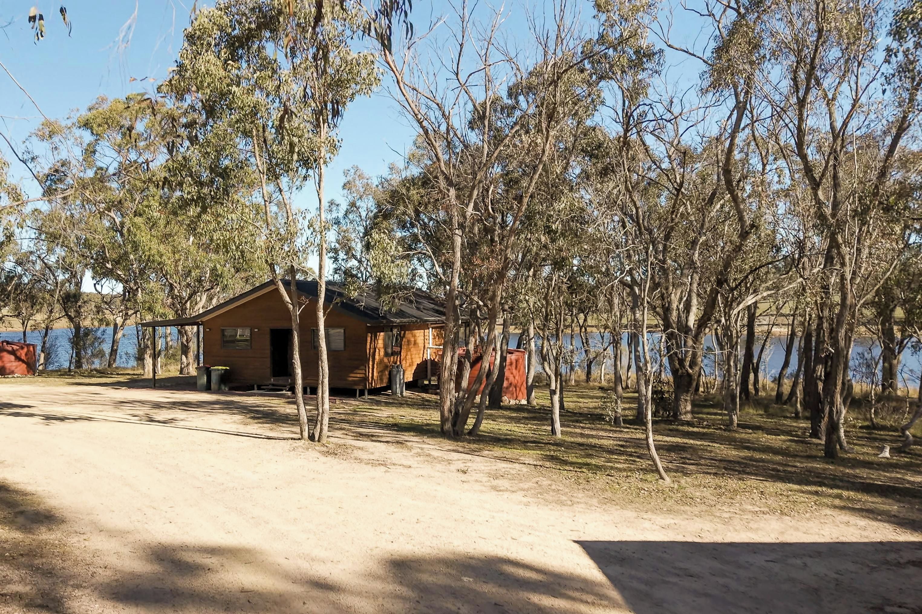 A cabin with a dirt ground, surrounded by trees, near a body of water.