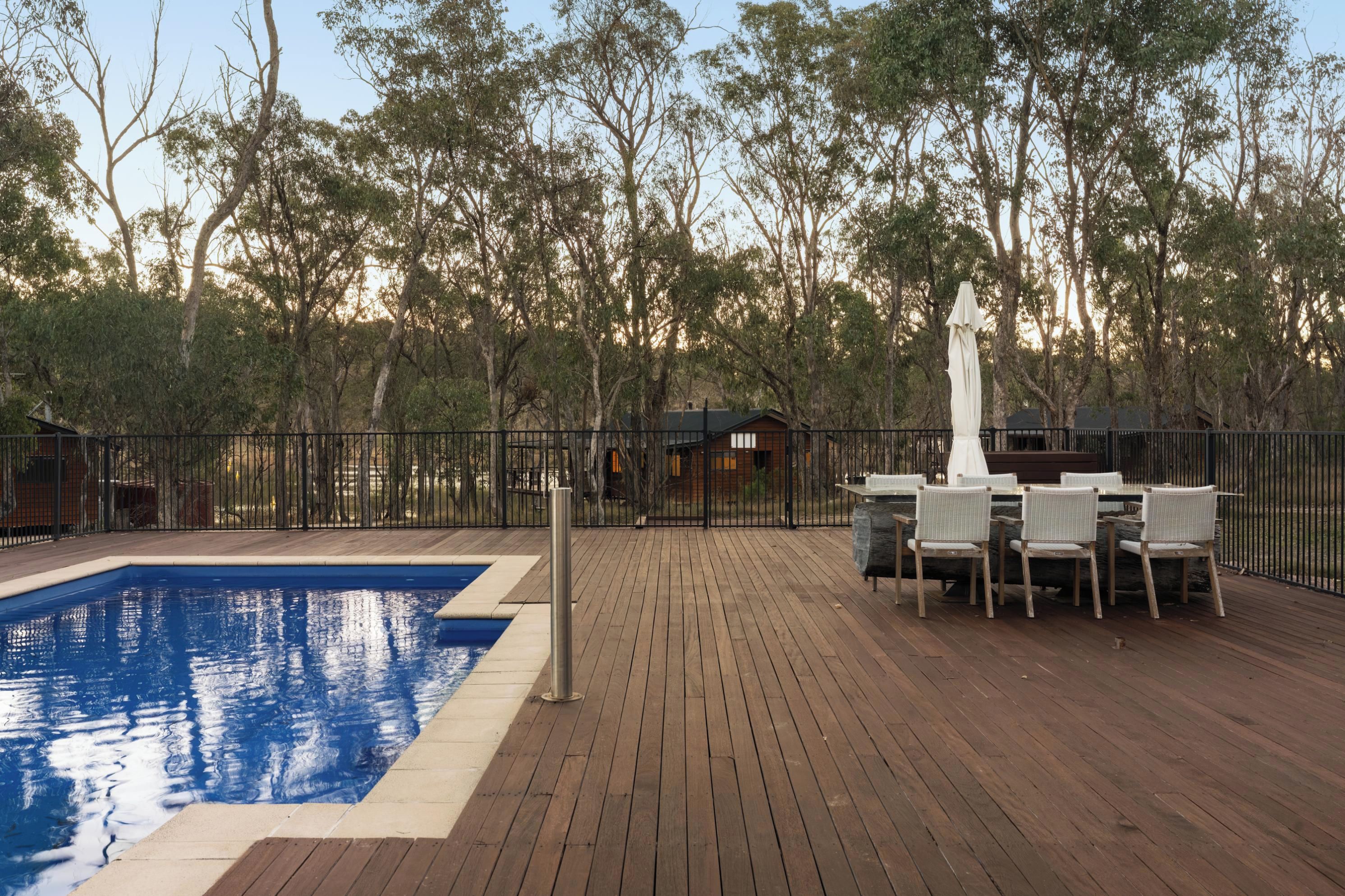 Una terraza de madera con piscina, rodeada de una valla y árboles.