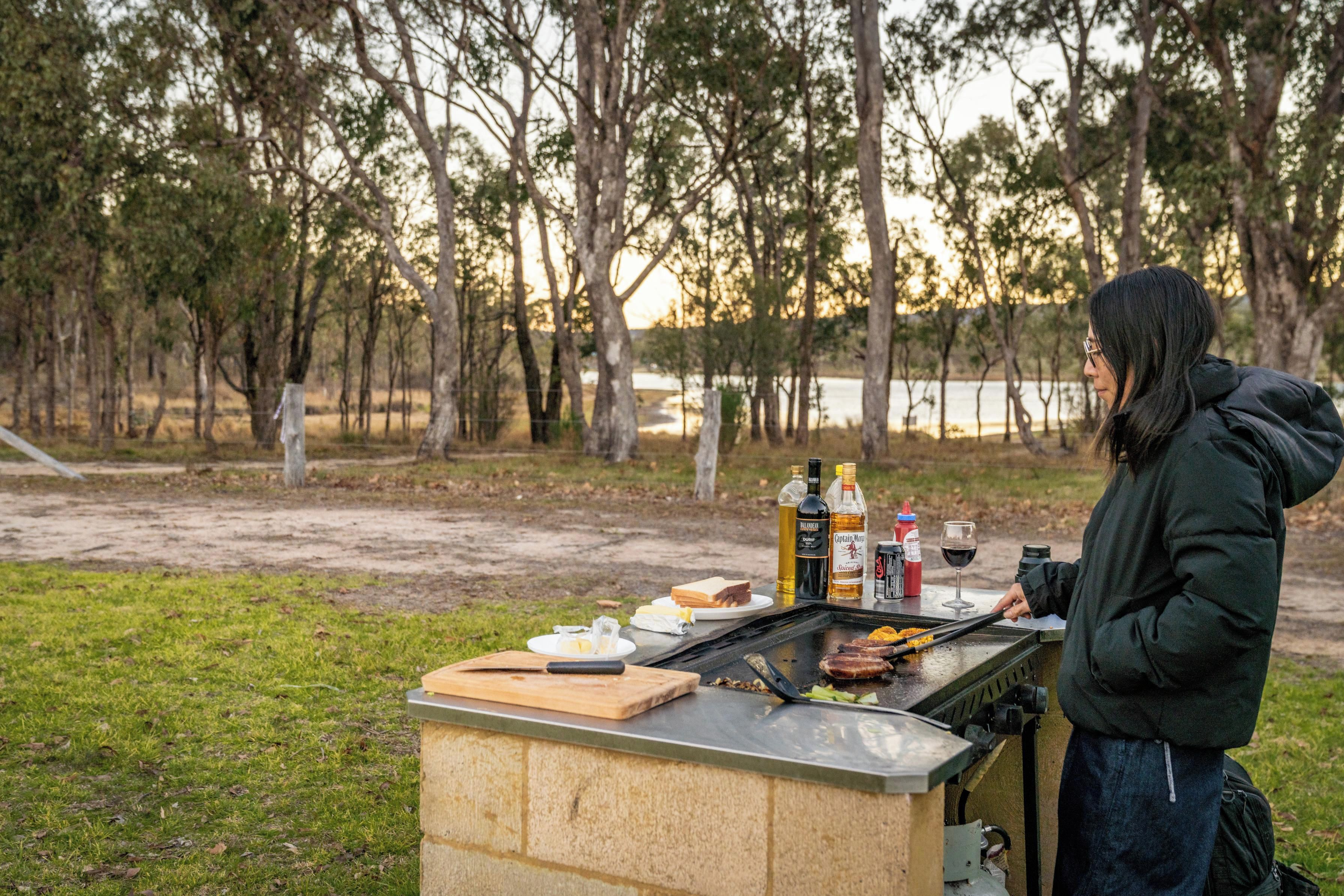An outdoor barbecue setup with a grill, food items, and beverages on a portable table.