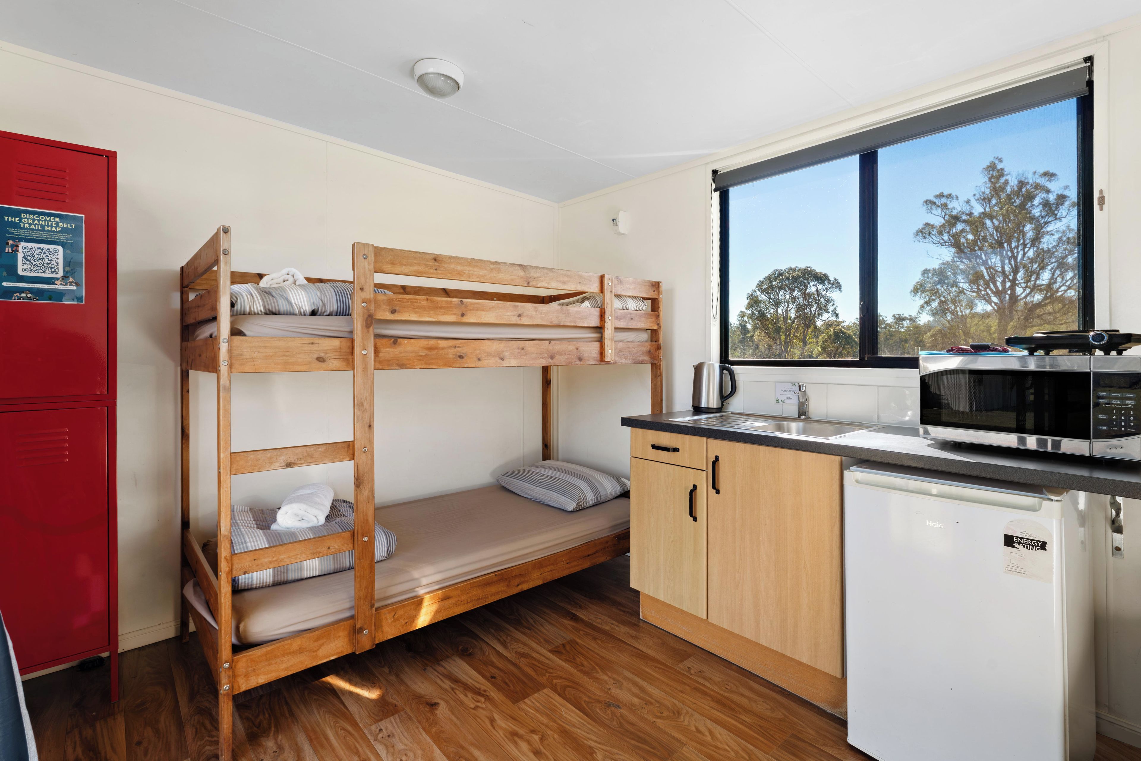 Een kleine kamer met een stapelbed, een kitchenette, een magnetron en een raam met uitzicht op bomen.