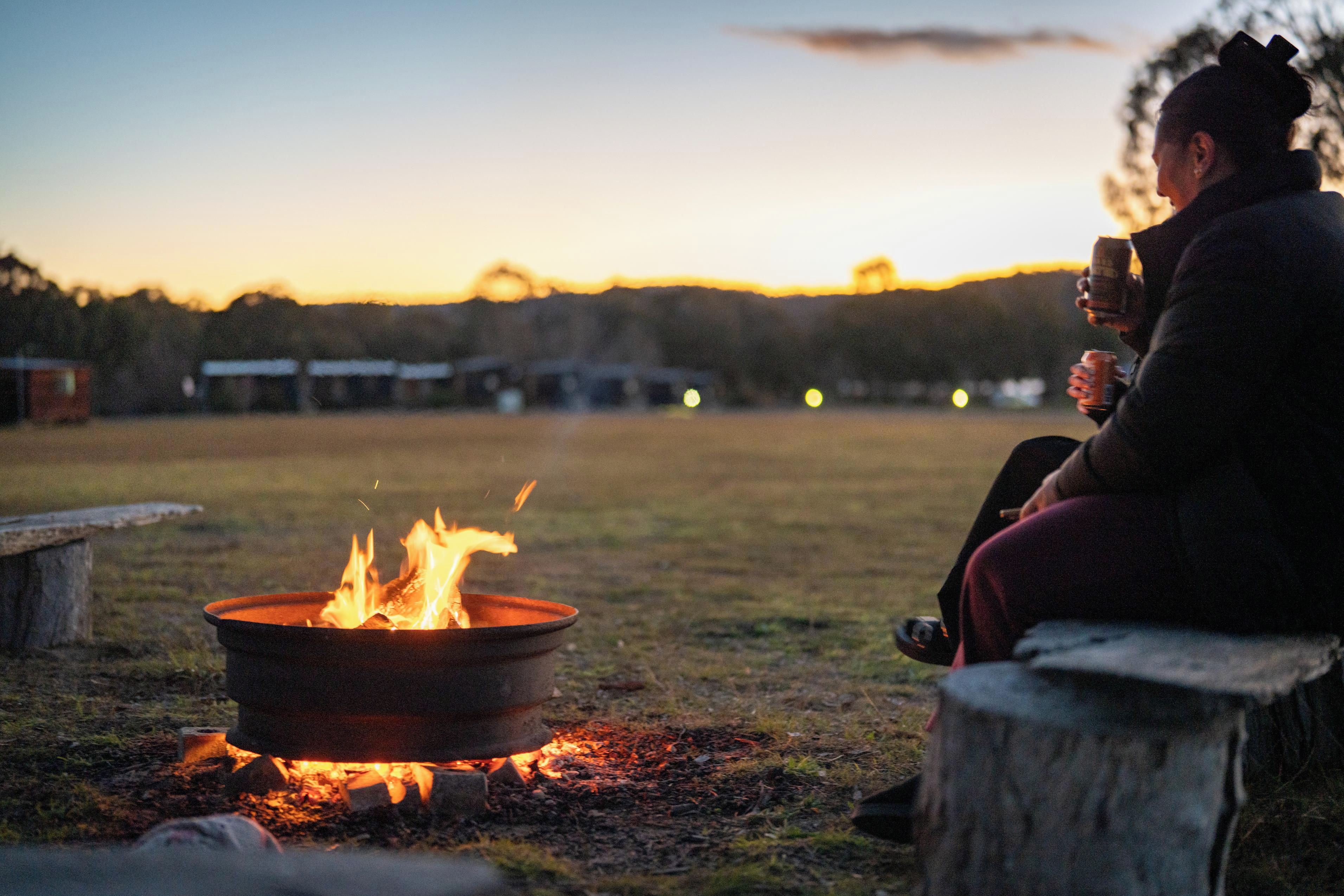 A person sitting by a campfire in a grassy field with a sunset in the background.