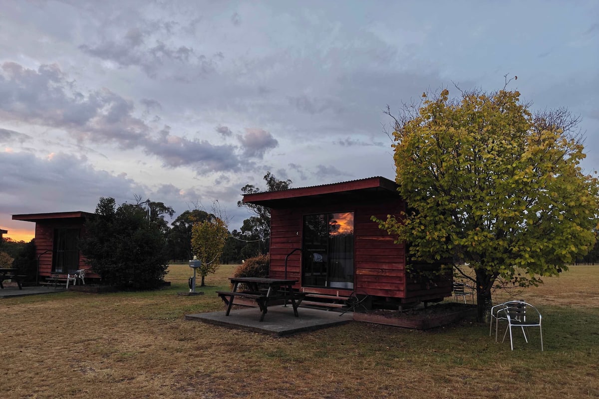 En enkel stuga med ett picknickbord och stolar på en gräsyta, omgiven av träd och en molnig himmel.