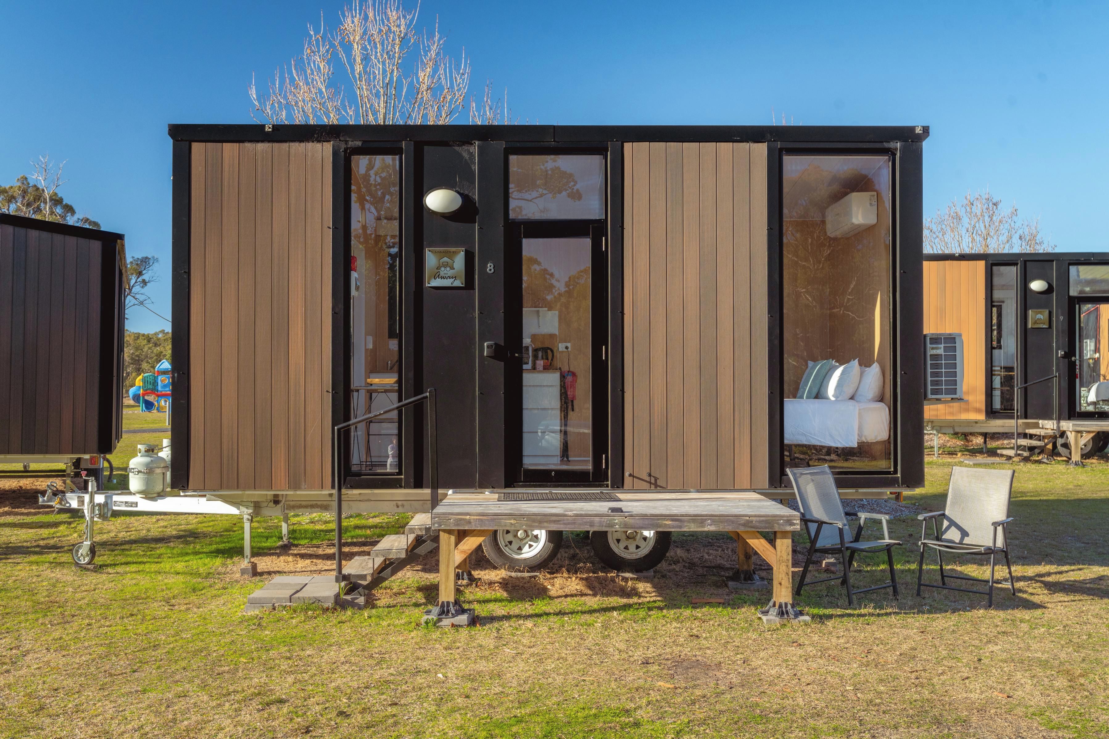 Una casa móvil moderna y compacta con un amplio ventanal corredera, un pequeño porche con mesa y sillas, y una cama visible en el interior.