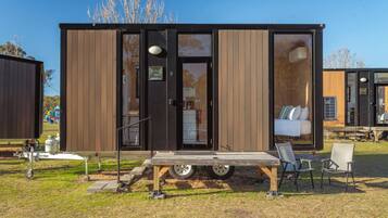 Una casa móvil moderna y compacta con un amplio ventanal corredera, un pequeño porche con mesa y sillas, y una cama visible en el interior.