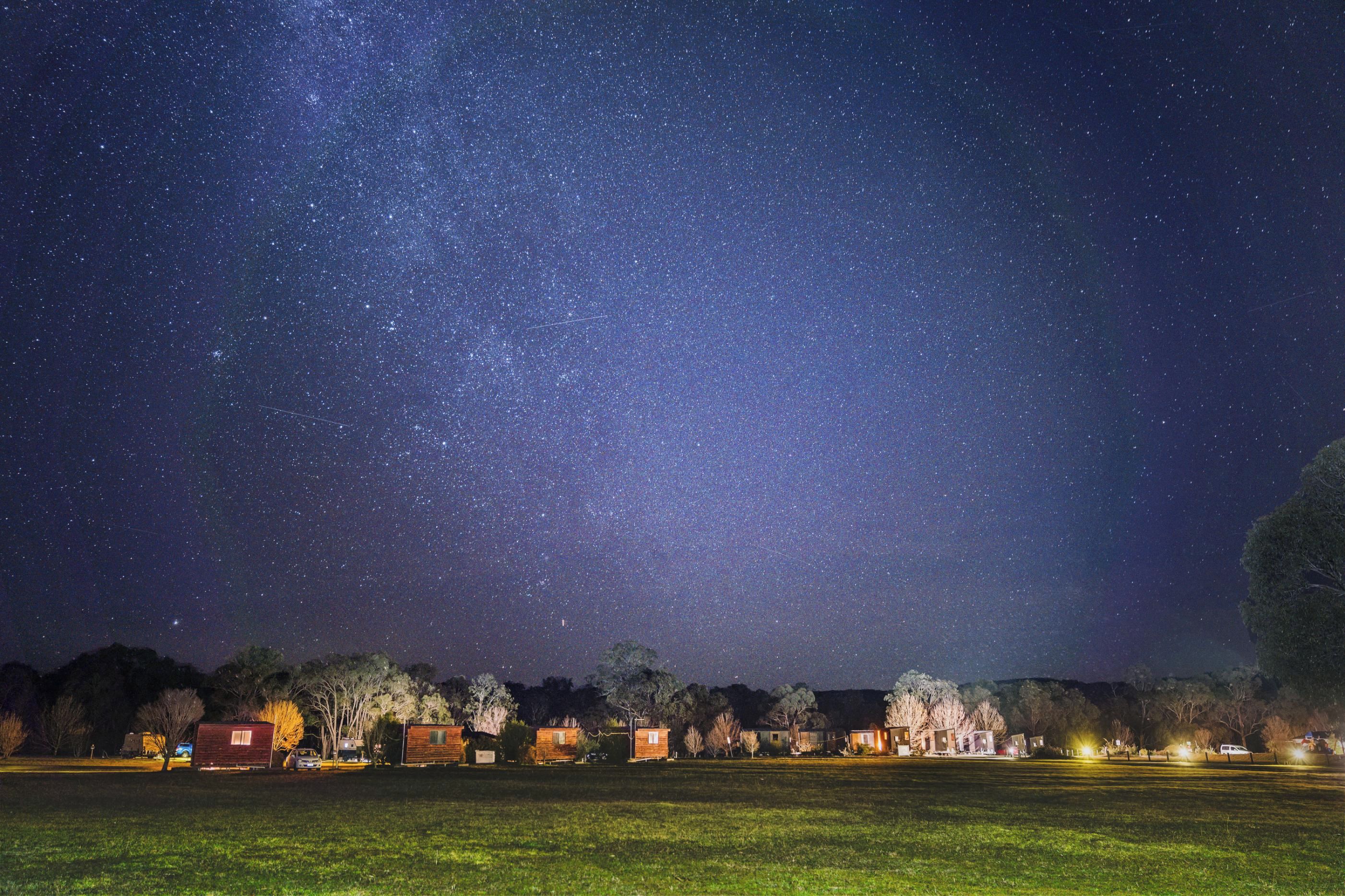 A starry night sky over a grassy field with several wooden cabins and a small building with lights on.