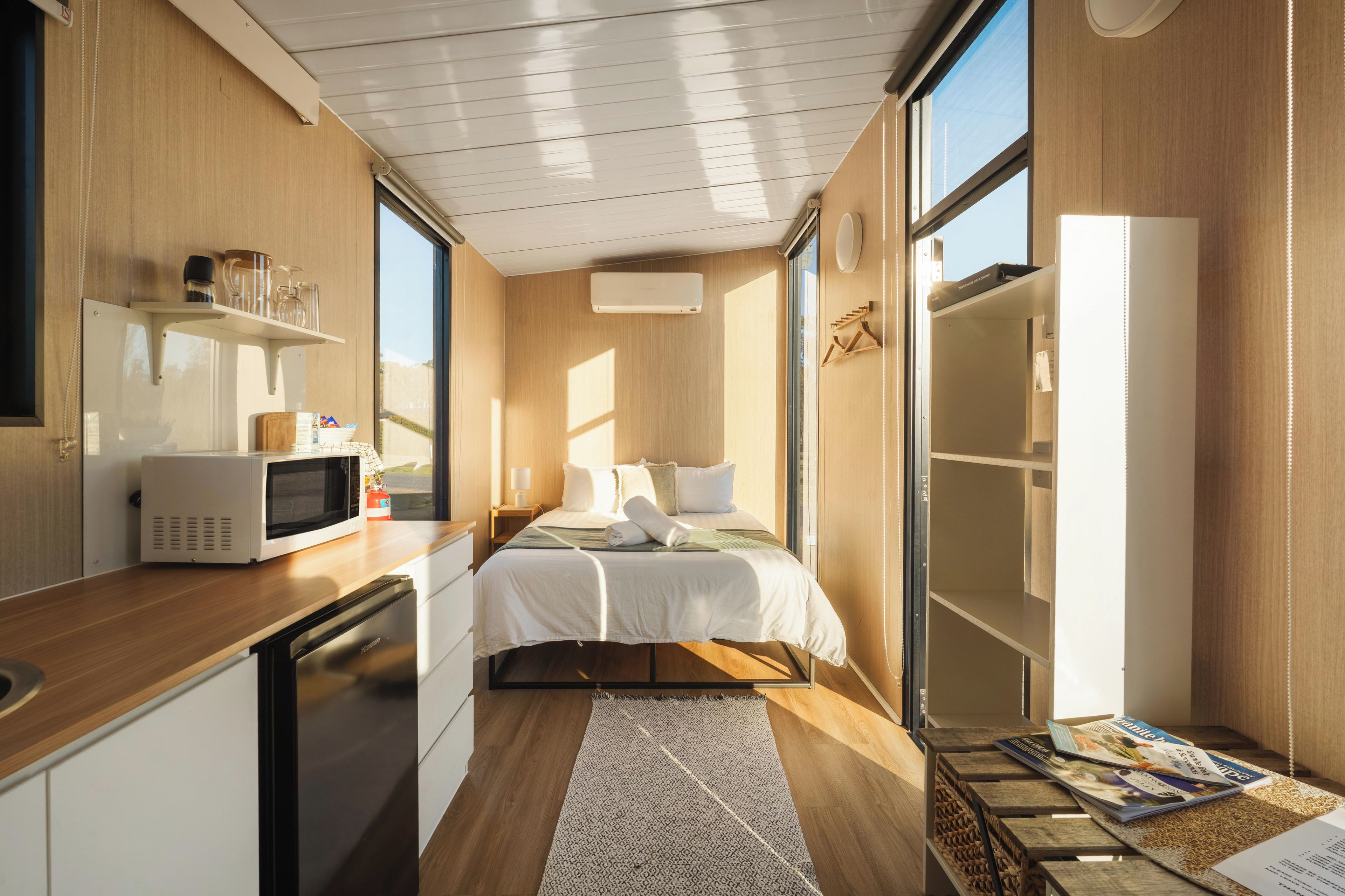 Une chambre d’hôtel compacte, dotée d’une kitchenette, d’un lit avec une literie blanche, d’une grande fenêtre et d’un plancher en bois.