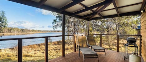 A covered outdoor deck with a view of a lake, two lounge chairs, and a barbecue grill.