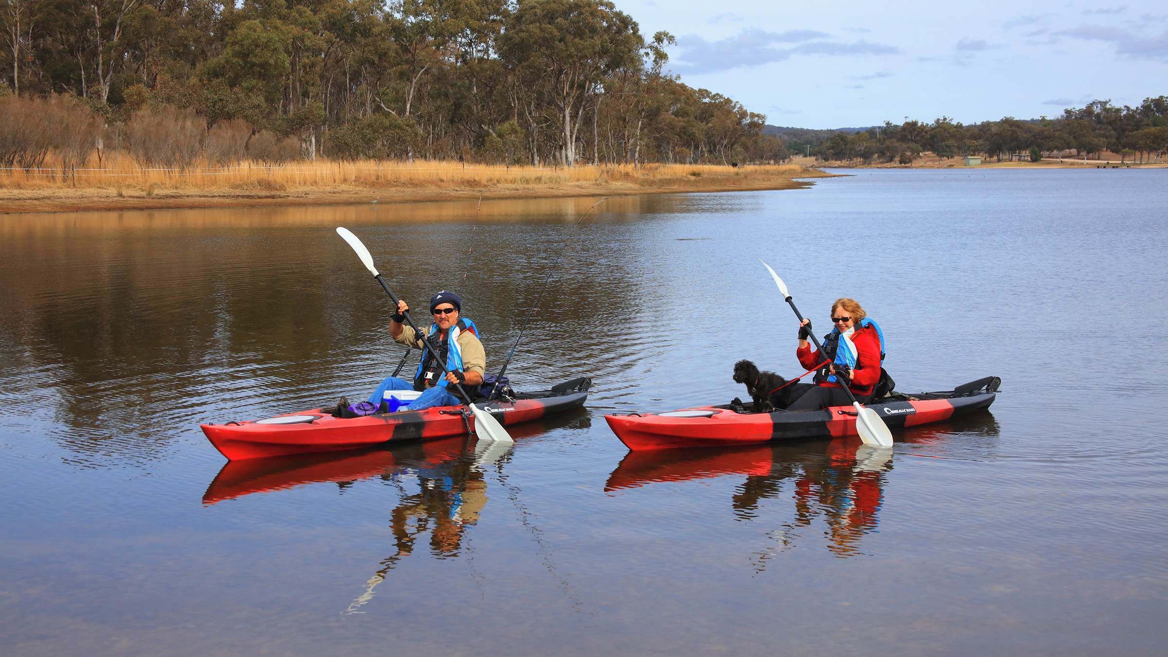 Two people kayaking on a calm lake with trees in the background.