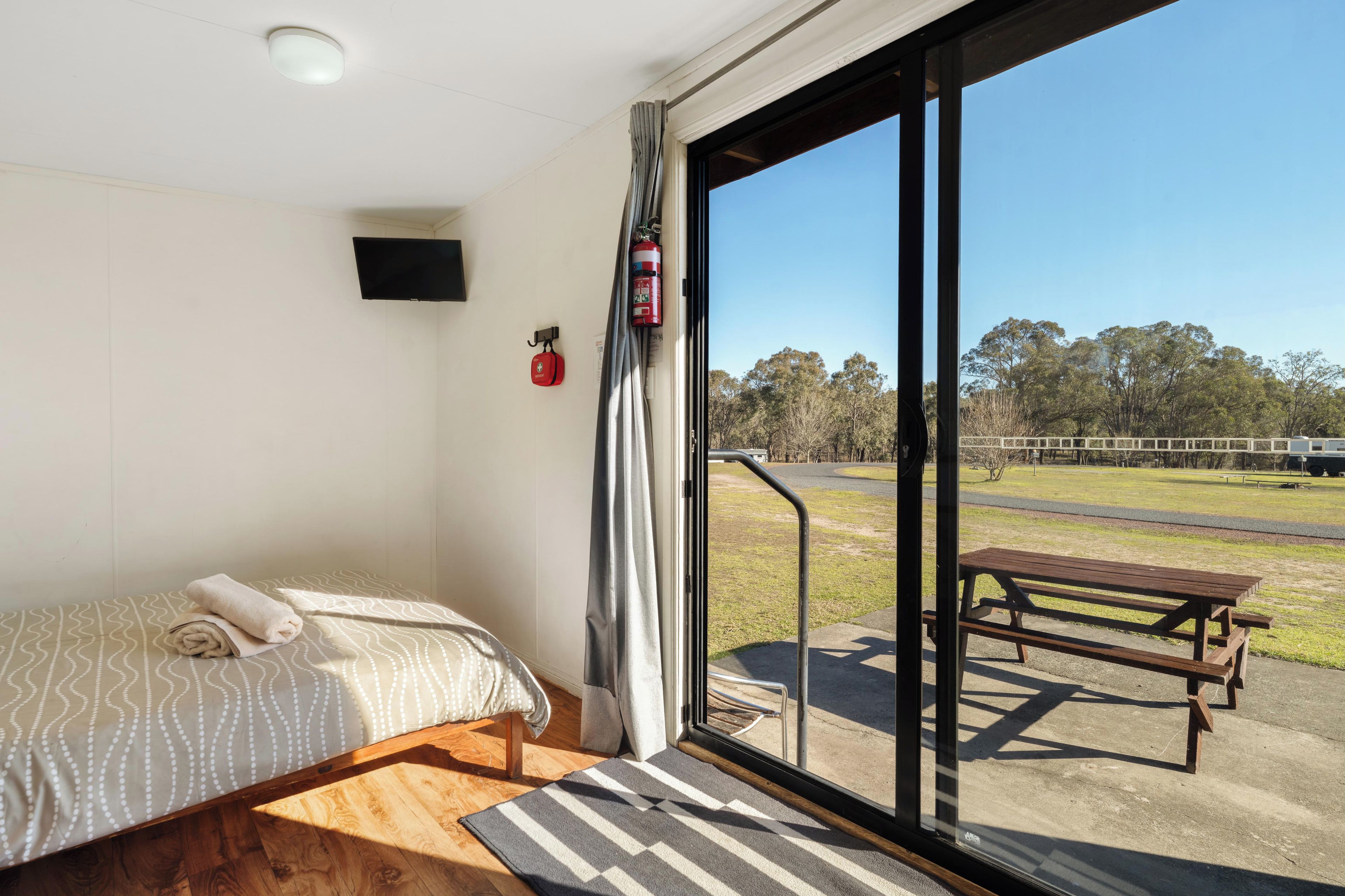 A room with a bed, a fire extinguisher, and a view of a park through a glass door.