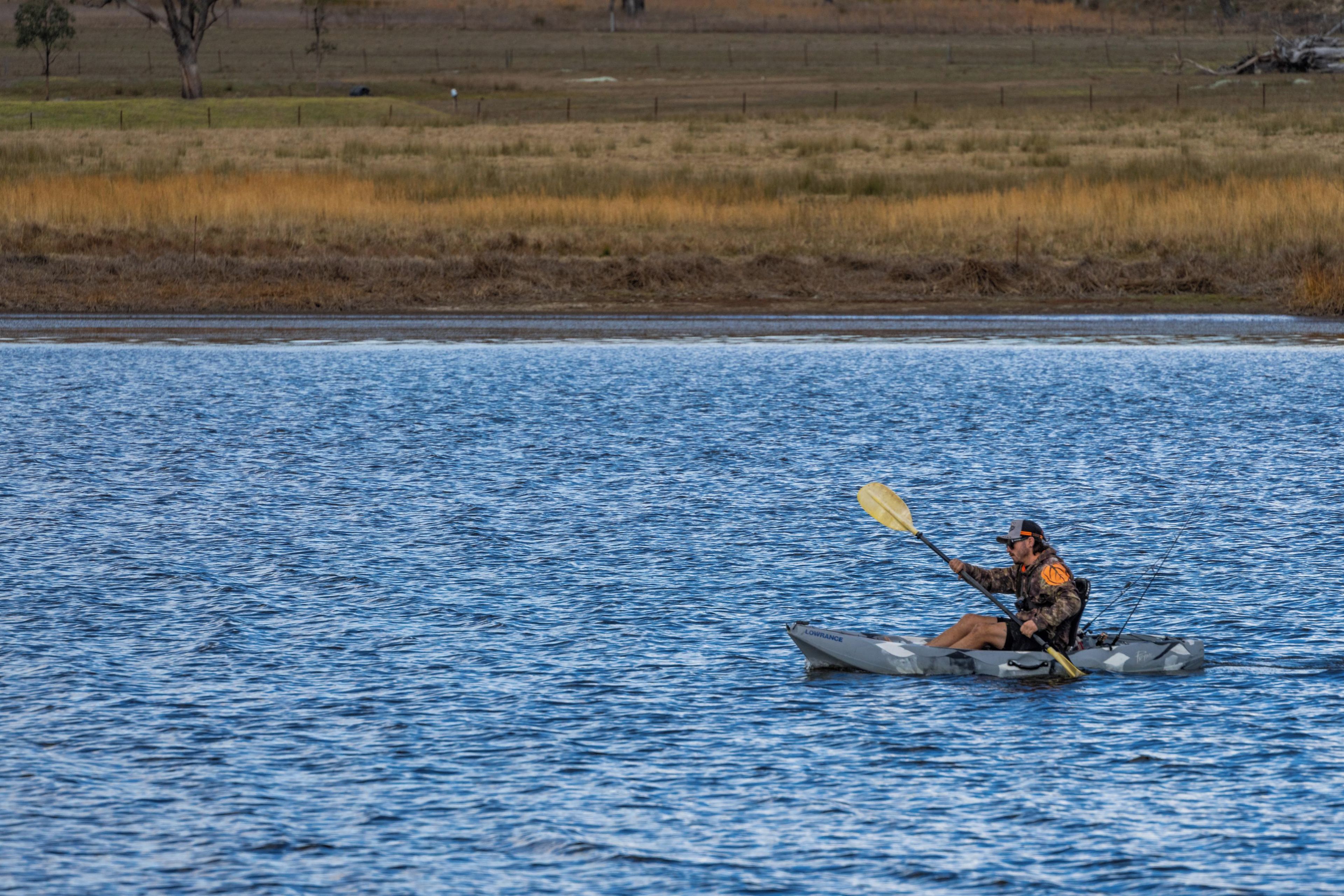 A person kayaking on a body of water with a grassy field in the background.
