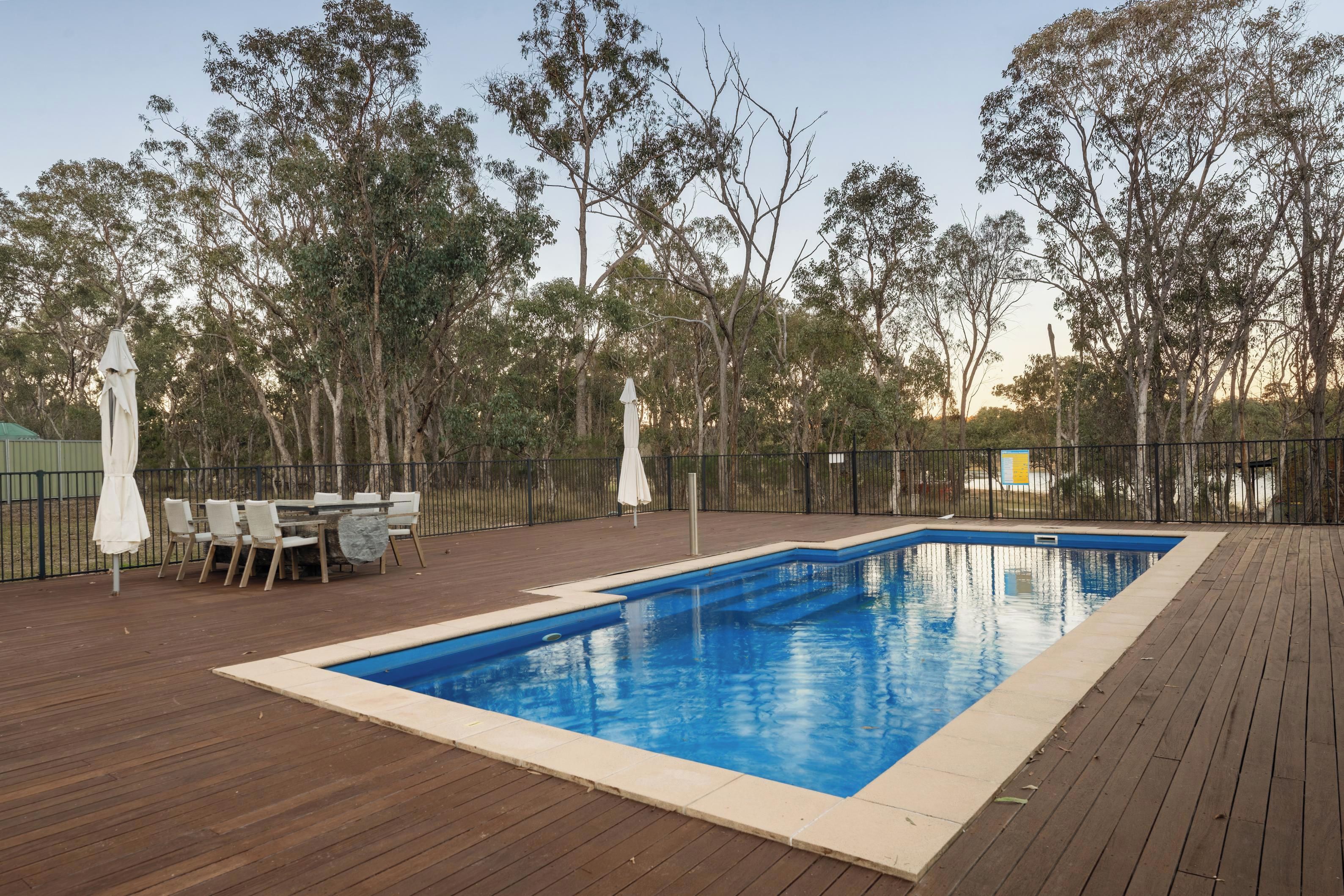 A rectangular pool with a wooden deck surrounding it, a table with chairs, and umbrellas.