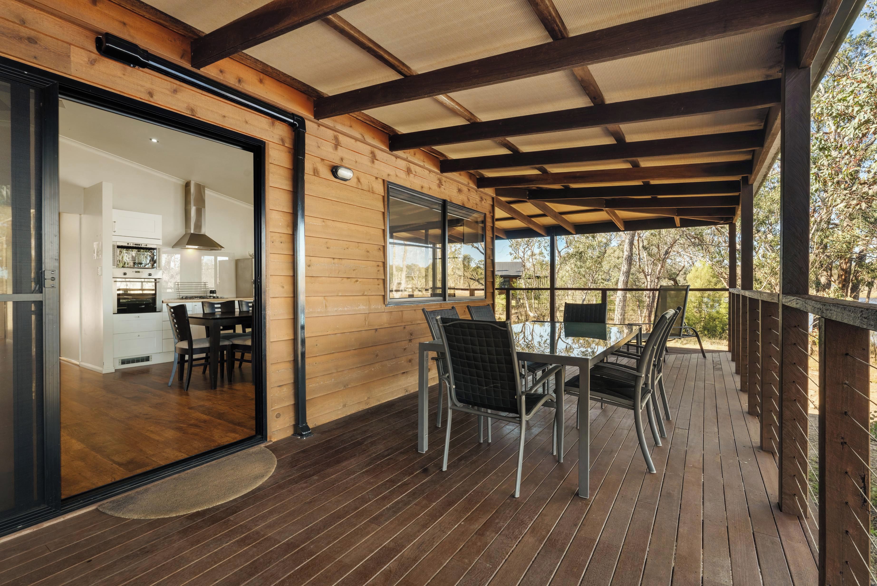 Terraza de madera con comedor y cocina visibles a través de un ventanal.