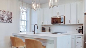 Cocina moderna con armarios blancos, una isla central con taburetes altos, iluminación colgante y un ventanal grande.