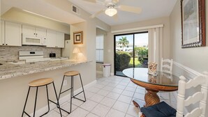 A modern kitchen with a granite countertop, white cabinets, a microwave, and a stove. There are two bar stools and a dining table with chairs. A ceiling fan and a window with blinds are visible. - San marco villas 2-bedroom newly updated condo in lovely Marco Island  (Marco Island)