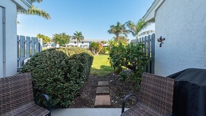 A patio area with two wicker chairs, a small garden with a hedge and a tree, and a view of a residential area with houses and palm trees. - San marco villas 2-bedroom newly updated condo in lovely Marco Island  (Marco Island)