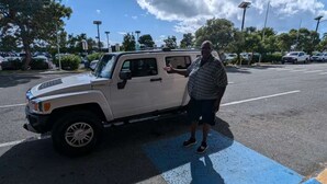 A person standing next to a white SUV in a parking lot with trees and other vehicles in the background.