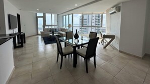 A spacious living area with a dining table, chairs, a sofa, and a TV. Large windows offer a view of the city. - Almendra I Spacious 1 Bedroom With Balcony (Panama City)