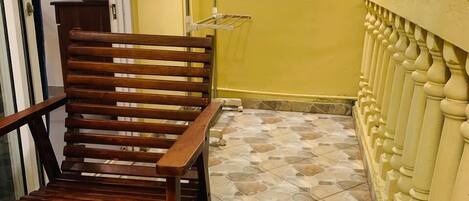 Un banco de madera y una silla en un balcón con suelo de baldosas, una pared amarilla y una lámpara.