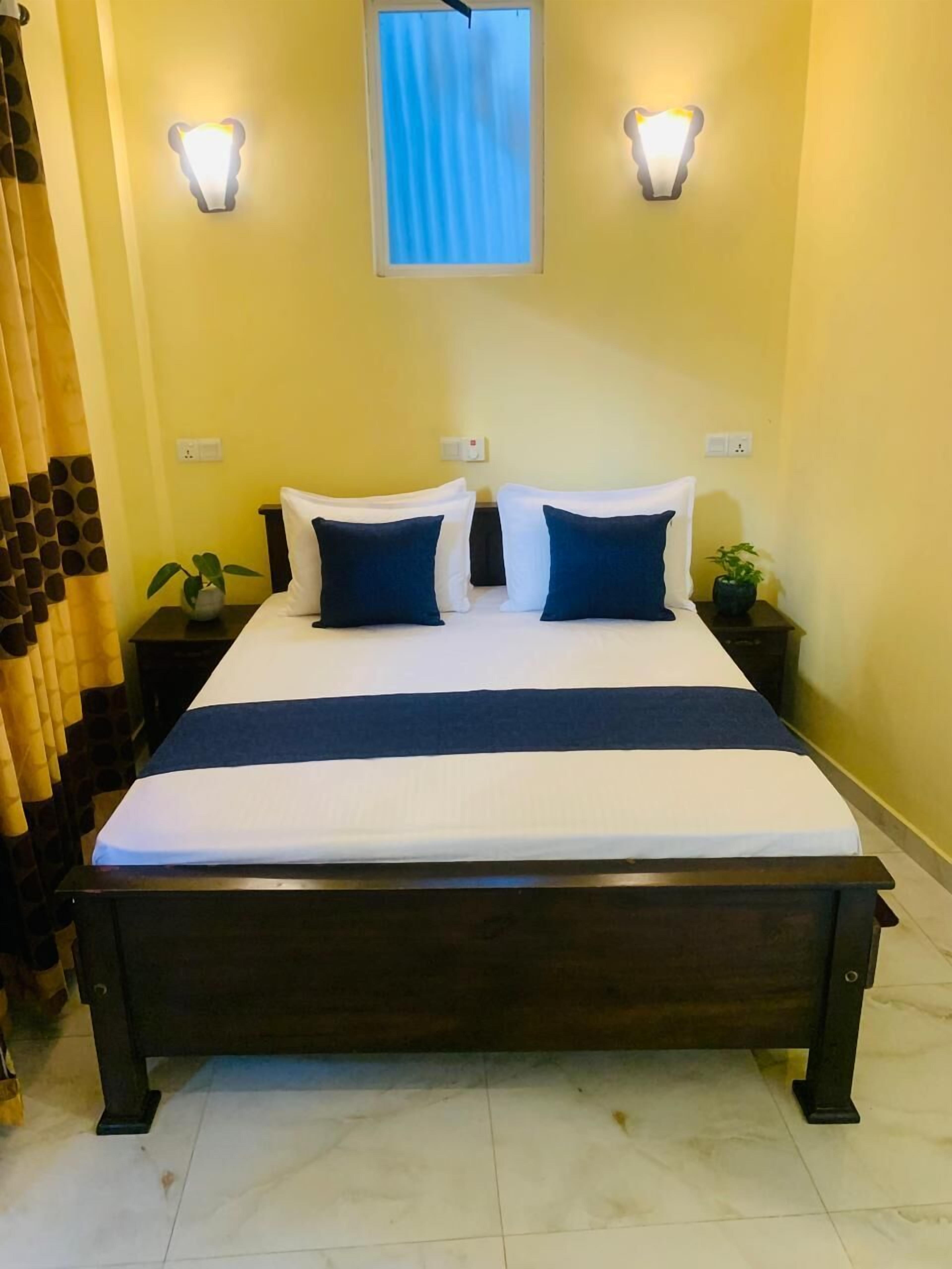 Una cama con ropa de cama blanca y azul, dos mesitas de noche con lámparas, una ventana con cortinas y una planta sobre una de las mesas.