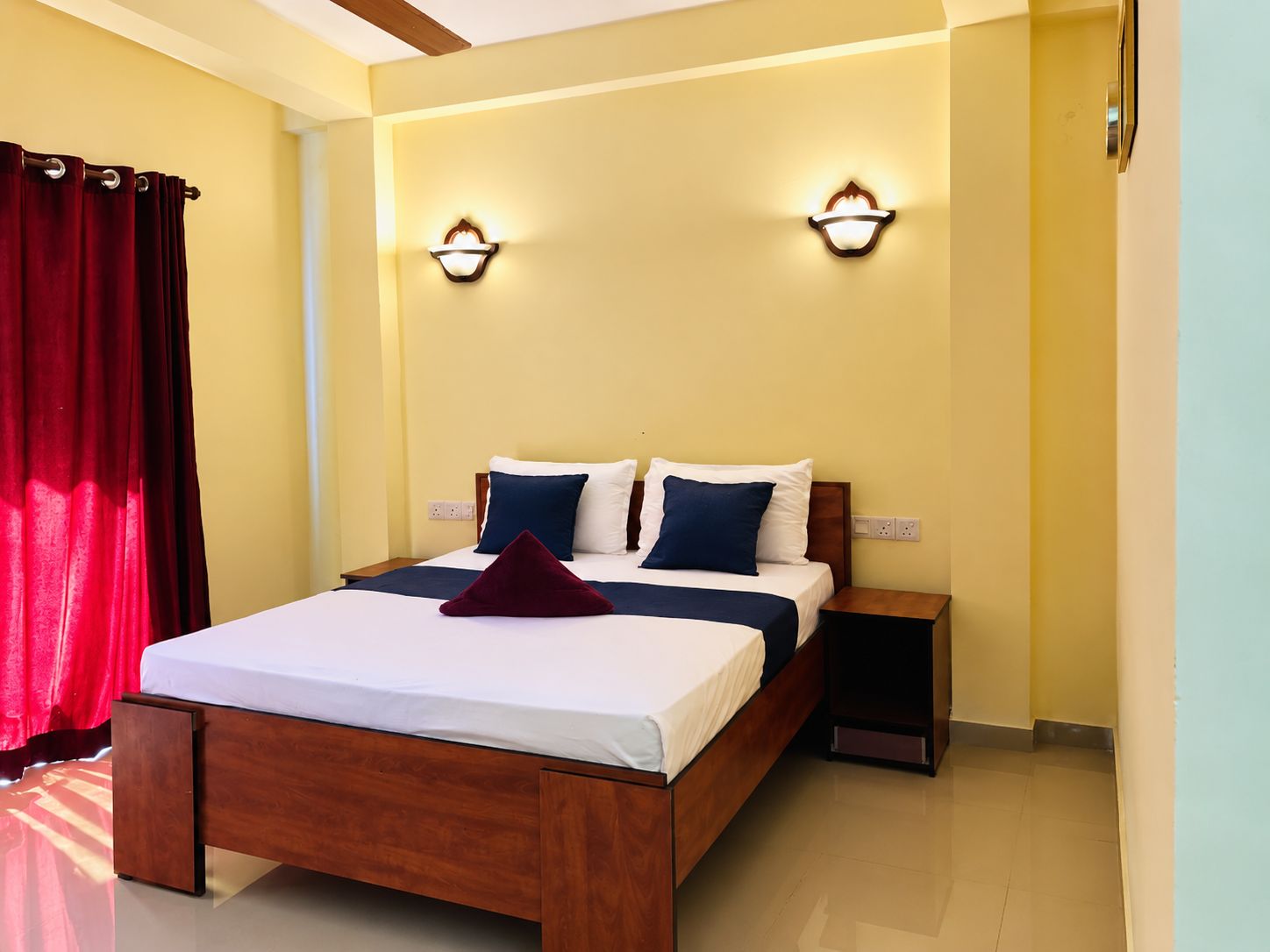 A hotel room with a wooden bed, two wall-mounted lights, a red curtain, and a nightstand.