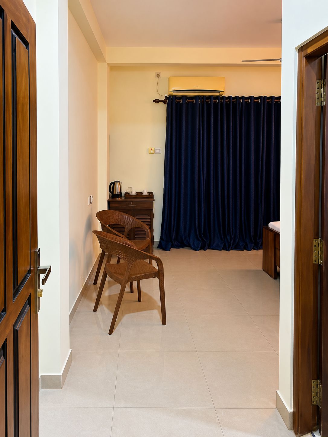 Una habitación de hotel con puerta de madera, una cama, una mesita con hervidor, una silla y una cortina azul.