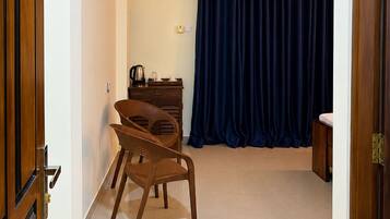 Una habitación de hotel con puerta de madera, una cama, una mesita con hervidor, una silla y una cortina azul.