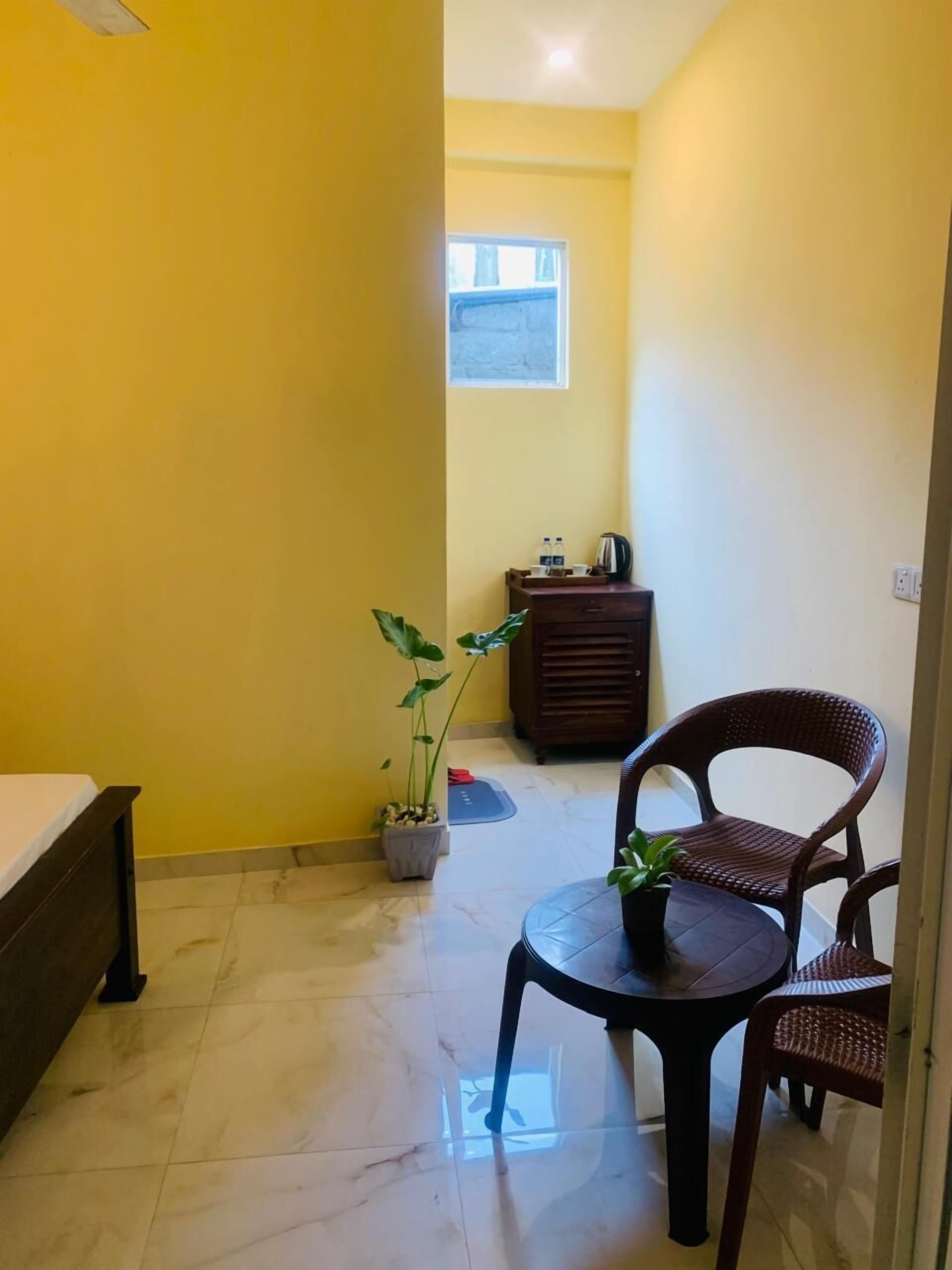 Una habitación pequeña, luminosa, con paredes amarillas, una cama, una mesita con una planta y una silla.