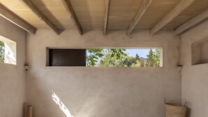 Una habitación con piso de concreto, ventana con vista a árboles, una cesta tejida y un tapete azul estampado.