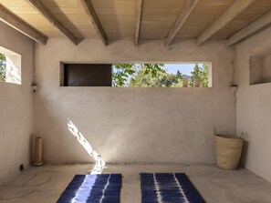 Ein Zimmer mit Betonboden, einem Fenster mit Blick auf Bäume, einem gewebten Korb und einer blauen, gemusterten Matte.