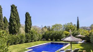 Una piscina rectangular con agua azul cristalina, rodeada de césped cuidado, árboles altos y algunas sombrillas de paja.
