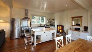Un'ampia cucina con pavimento in legno, mobili bianchi, un'isola centrale con sgabelli, un frigorifero in acciaio inossidabile, un piano cottura e un camino.