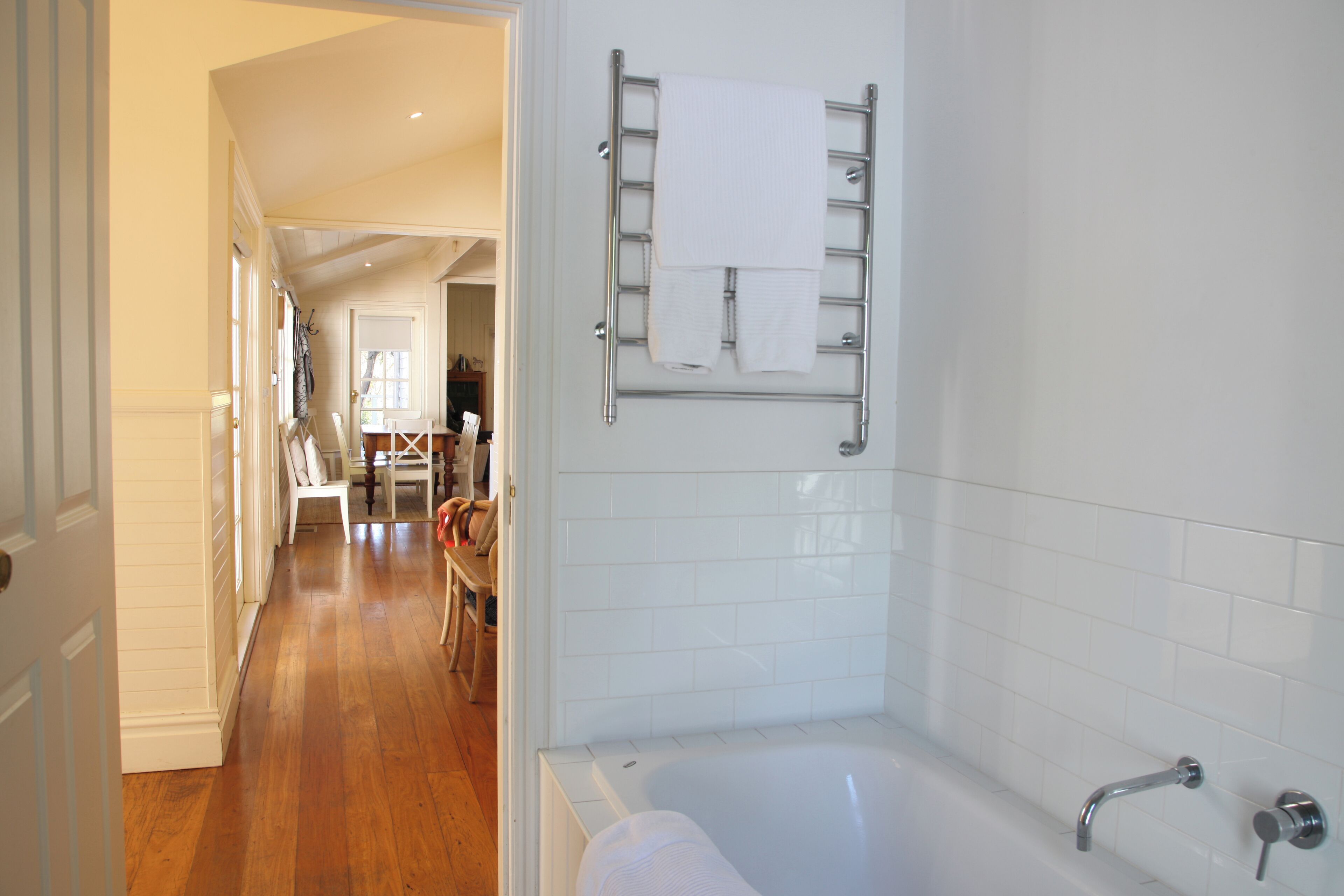 A bathroom with a bathtub, a towel rack, and a wooden floor leading to a dining area.