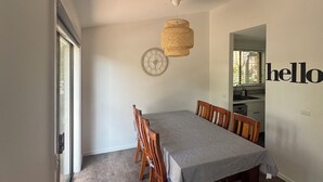 Zona pranzo con un tavolo lungo, sedie di legno, un lampadario intrecciato e vista sulla cucina attraverso un'apertura.