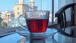Une tasse de thé en verre posée sur une table en verre transparent, avec en arrière-plan la vue d’un bâtiment doté de deux minarets.
