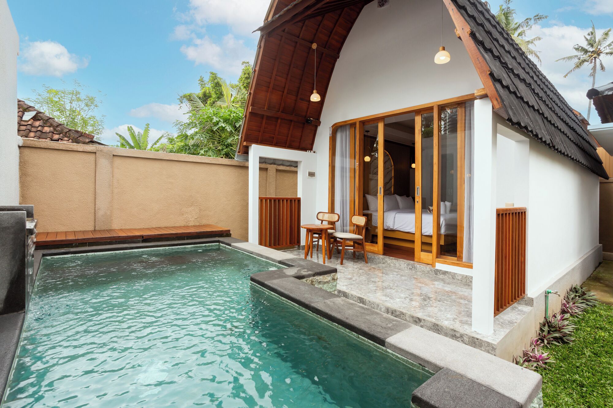 Un área junto a la piscina con una terraza de madera, una pequeña mesa con sillas, y una habitación con cama y ventana.